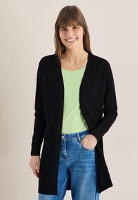 Femme souriante aux cheveux bruns lisses portant un cardigan noir, un haut vert clair et un jean bleu, debout avec une main dans la poche contre un mur uni.