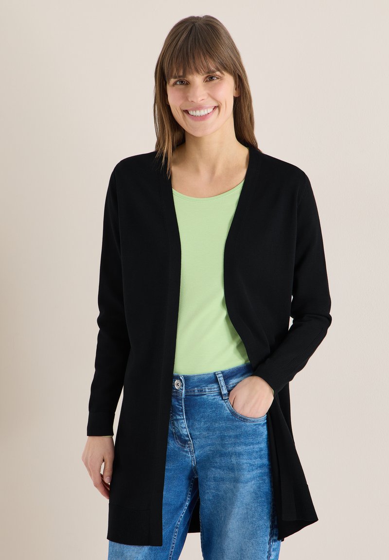 Femme souriante aux cheveux bruns lisses portant un cardigan noir, un haut vert clair et un jean bleu, debout avec une main dans la poche contre un mur uni.