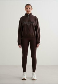 Sudadera marrón con cremallera y el texto "ALTN' SPORTSWEAR", combinada con leggings marrones a juego y zapatillas blancas, sobre un fondo neutro.