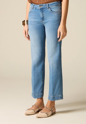 Žena nosící světle modré džíny s rovnými nohavicemi, hnědý vzorovaný top, béžové špičaté baleríny s pásky se vzhledem hadí kůže a zlatý náramek manžeta.