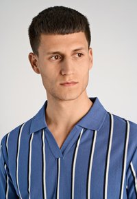 Niebieska koszulka polo w paski z pionowymi białymi i czarnymi paskami, z dekoltem w kształcie litery V, krótkimi rękawami oraz teksturowanym materiałem.