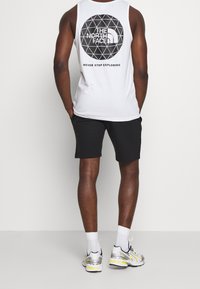 Bílý bezrukávový top s geometrickým černým logem na zadní straně, v kombinaci s černými šortkami a šedými teniskami se žlutými akcenty.