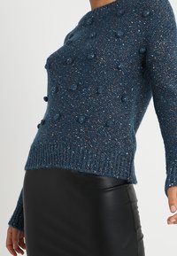 Pull bleu en maille avec des boules texturées, un col rond et des poignets côtelés, associé à une jupe en cuir noir.