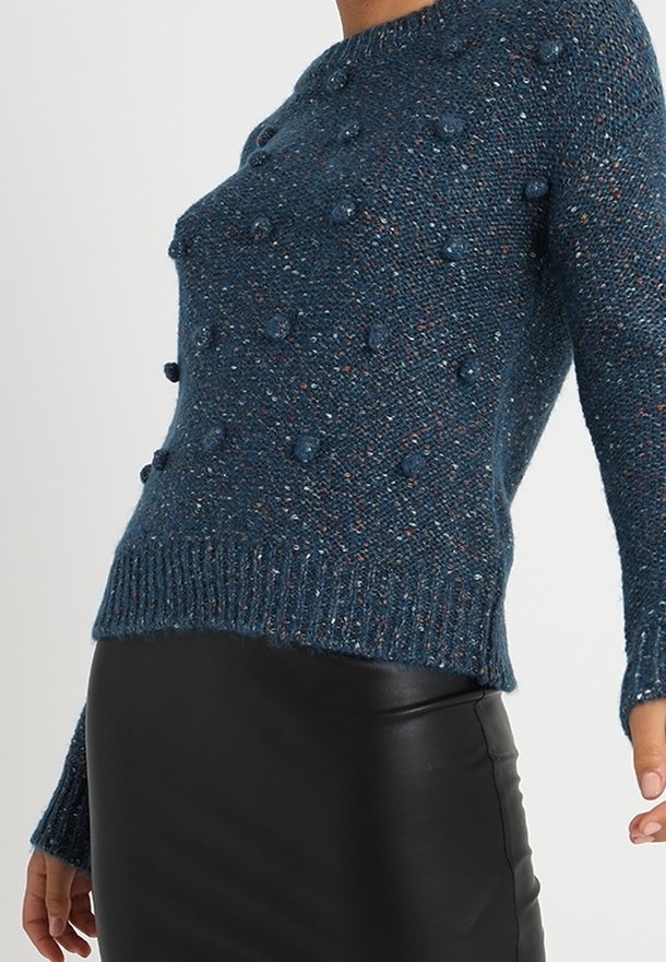 Pull bleu en maille avec des boules texturées, un col rond et des poignets côtelés, associé à une jupe en cuir noir.