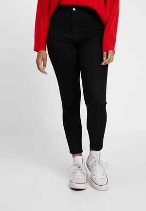 Person iført sorte højtaljede skinny bukser, hvide højskaftede sneakers og en rød langærmet sweater, stående på en ensfarvet baggrund.
