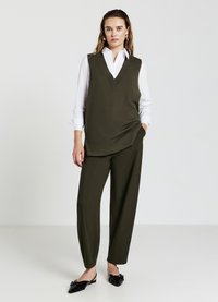 Top senza maniche di colore verde oliva con scollatura a V, indossato sopra una camicia bianca a bottoni. Pantaloni verde oliva di taglio comodo e scarpe nere a punta.