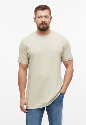 Man met kort haar en baard, draagt een beige shirt met korte mouwen en blauwe spijkerbroek, staand met één hand in de zak tegen een witte achtergrond.