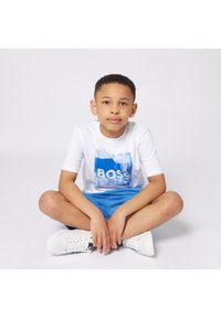 T-shirt blanc avec un graphique bleu "BOSS", associé à un short bleu clair. Le garçon est assis en tailleur, portant des baskets blanches. Fond uni.