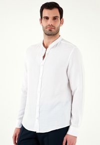 Camisa blanca de manga larga con cuello mao, tejido texturizado y cierres de botones en la parte delantera. Combinada con pantalones oscuros.