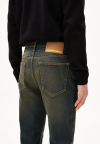 Indigo denim jeans met een licht vervaagde afwerking, voorzien van een bruine leren label met de tekst "ARMEDANGELS" en contrasterende stikseldetails.