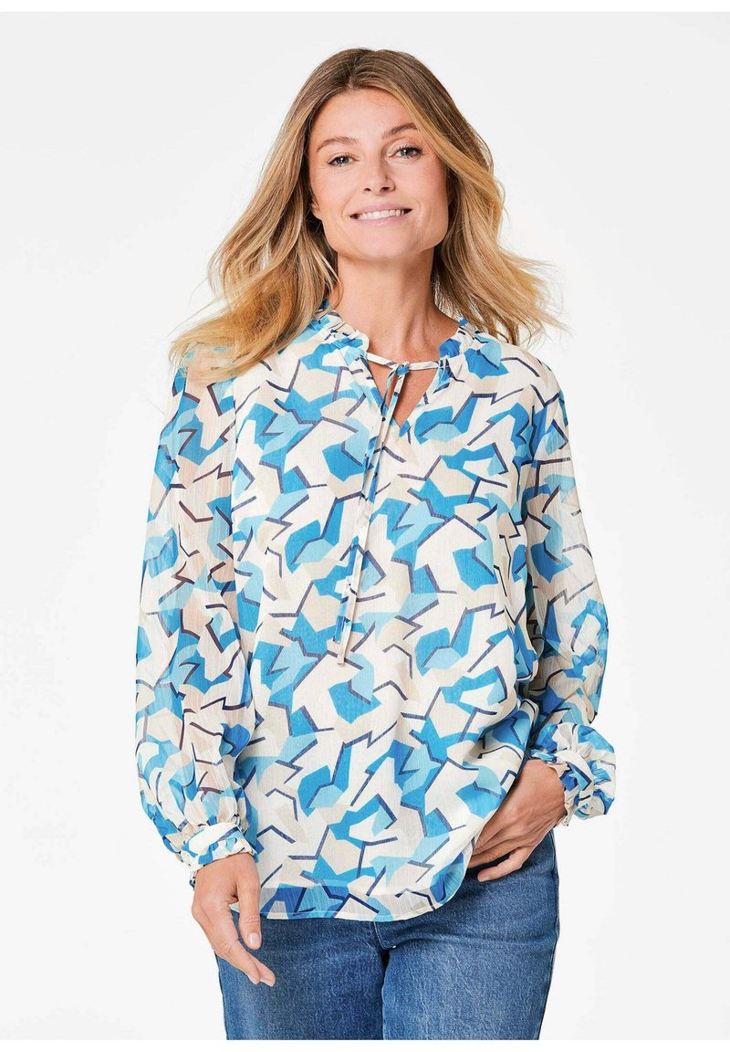 Langärmlige Bluse aus leichtem Stoff mit einem blau-weißen geometrischen Muster, Rundhalsausschnitt und Bindedetail, kombiniert mit Jeans.