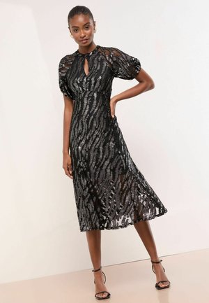 Žena stoji sa rukom na bokovima noseći crnu, prozirnu midi haljinu s nahuškanim rukavima i srebrnim šljokičastim prugama, uparenu s crnim cipelama s remenčićem oko članka.