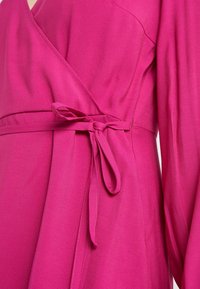 Różowa sukienka kopertowa związywana w talii, z długimi rękawami i gładką fakturą. Materiał pięknie się układa, eksponując jednolity kolor.