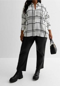 Blusa bianca con motivo a quadri neri, colletto e tasca abbinata a pantaloni neri a gamba larga e stivali neri con tacco largo, mentre tiene una borsa trapuntata.