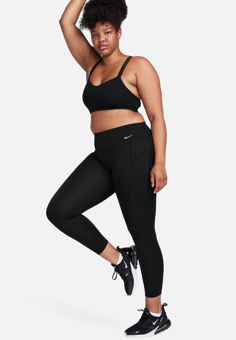 Conjunto deportivo negro que incluye un sujetador deportivo ajustado y corto, además de leggings de tiro alto con un logo. El modelo lleva zapatillas negras.