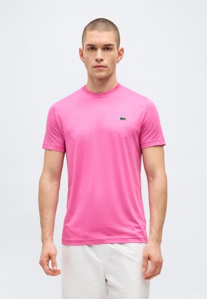 Man with short hair wearing a bright pink short-sleeve shirt and white pants, standing against a plain light background.