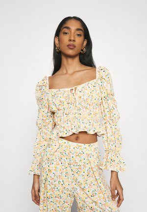 Top floral curto com mangas bufantes, detalhes franzidos, decote quadrado e barra com volantes. Padrões amarelos, laranjas e verdes sobre um fundo branco.