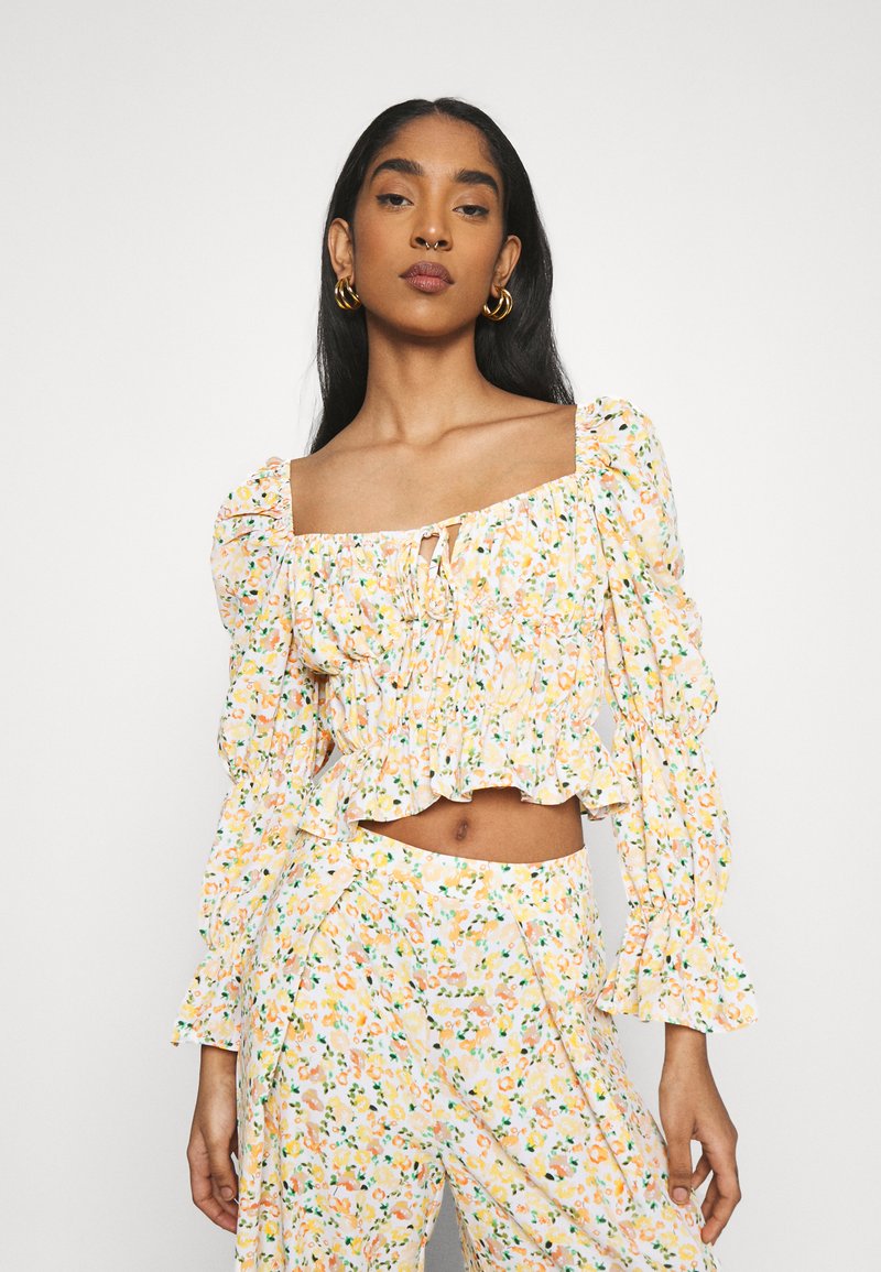 Top floral curto com mangas bufantes, detalhes franzidos, decote quadrado e barra com volantes. Padrões amarelos, laranjas e verdes sobre um fundo branco.