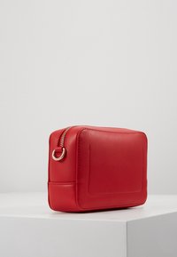 Mała prostokątna czerwona skórzana saszetka z złotym zamkiem błyskawicznym i detalem w postaci pierścienia, położona na białej powierzchni na tle jasnoszarym.