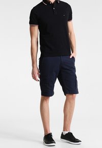 Uomo che indossa una polo nera, pantaloni cargo blu navy, calze bianche e sneakers nere, in piedi con una mano in tasca.
