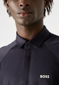 Camisa abotoada escura em azul-marinho com colarinho clássico, apresentando o logótipo branco "BOSS" no lado esquerdo do peito e uma textura suave.