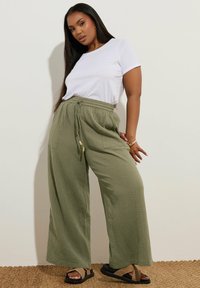 Celana panjang lebar berwarna hijau zaitun dengan bahan tekstur, pinggang elastis, dan tali pengikat. Dipadukan dengan kaos putih lengan pendek dan sandal hitam.