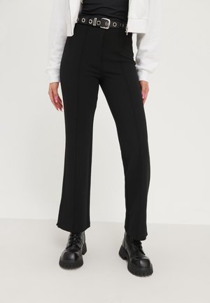Personne portant un pantalon noir taille haute avec une ceinture noire et des bottes noires à lacets, associé à une veste blanche et un haut noir.