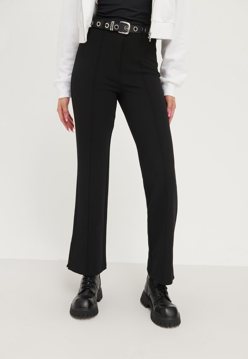 Personne portant un pantalon noir taille haute avec une ceinture noire et des bottes noires à lacets, associé à une veste blanche et un haut noir.
