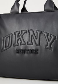 Melnas ādas iepirkumu soma ar reljefu izgrieztām "DKNY" un "NEW YORK" logo, apaļām rokturiem un gludu virsmu. Minimalistisks dizains ar kontrasta šuvēm.