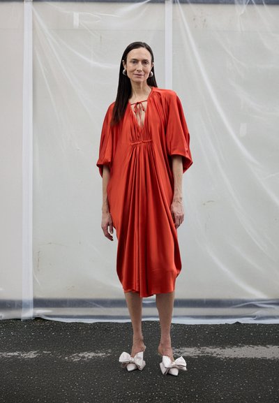 Vestido rojo fluido con un escote profundo y detalles fruncidos en el corpiño. Combinado con zapatos blancos que tienen grandes lazos. Textura de tela suave.