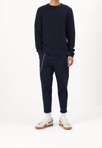 Námořnicky modrý svetr z texturovaného úpletu, volné námořnicky modré kalhoty s detailním řetízkem a béžové tenisky s černými akcenty a bílými ponožkami.