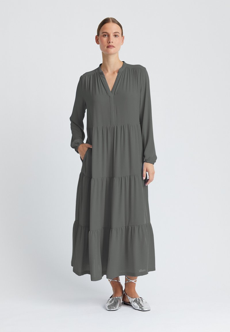 Robe longue à étages en tissu gris foncé, avec un décolleté en V, des manches longues et des poches latérales. Caractérisée par une texture légère et fluide.