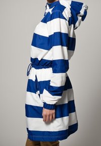 Blå och vit randig regnjacka med dragsko i midjan, huva och elastiska ärmslut. Tillverkad av ett vattentätt material och har en avslappnad passform.