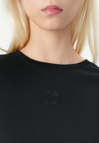 Black short-sleeve top with a crew neckline, featuring a subtle embossed logo in the center. Smooth texture with a fitted silhouette.