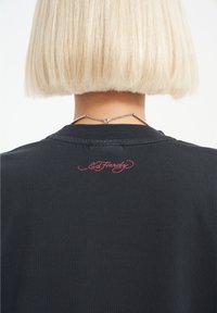 Svart t-shirt med rund halsringning, med en distinkt broderad rosa "Ed Hardy"-logotyp på baksidan nära kragen. Mjuk, slät textur.
