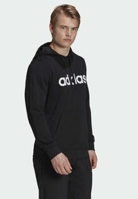 Felpa nera in cotone con un grande logo bianco "adidas" sul davanti, cappuccio regolabile con lacci e tasca a marsupio. Abbinata a pantaloni neri.