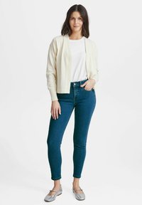 Béžový kardigan přes bílou košili, spárovaný s tmavě modrými skinny džíny a stříbrnými balerínami. Texturovaná látka, otevřený střih, přiléhavý tvar.