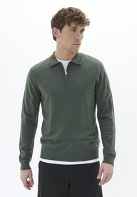 Zeleni pleteni pulover z ovratnikom na četrt zadrgo, dolgimi rokavi in prilegajočim se robom. Iz bombažne tkanine z gladko teksturo, položen preko belega spodnjega majice.