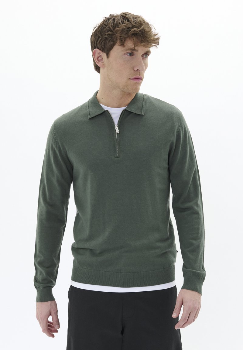 Zeleni pleteni pulover z ovratnikom na četrt zadrgo, dolgimi rokavi in prilegajočim se robom. Iz bombažne tkanine z gladko teksturo, položen preko belega spodnjega majice.