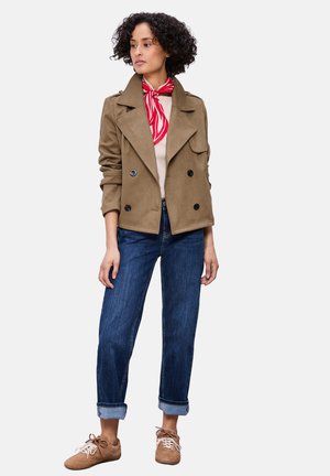 Femme aux cheveux bouclés portant une veste croisée beige, un foulard rouge à motifs autour du cou, un jean bleu retroussé et des baskets beiges, debout et regardant vers la gauche.