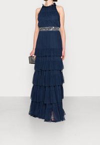 Marinblå maxiklänning i lager av genomskinlig tyll, med en volangdekorerad halsringning och pärlbroderi vid midjan. Modellen håller en liten svart kuvertväska.