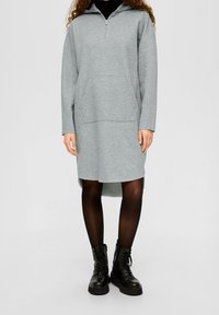 Šedé mikinové šaty s kapucí, s půlzipem vepředu, dlouhými rukávy a přední kapsou; doplněné o černé bojové boty a punčochy.