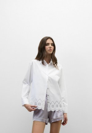 Jeune femme aux longs cheveux bruns porte une blouse blanche boutonnée avec un ourlet en dentelle festonnée et un short en satin gris clair devant un mur blanc uni.