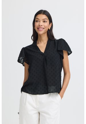 Naeratav naine kannab musta tekstuuriga pluusi volangiliste varrukatega ja valgeid pükse, seisab käed taskutes heleda tausta ees.