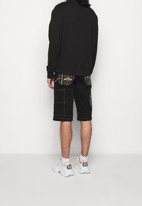 Black sweatshirt with long sleeves, paired with knee-length black shorts featuring patterned pockets and visible stitching details. White sneakers.
