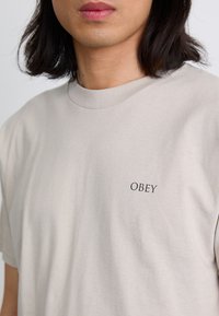 Világosszürke póló, pamutból készült, kerek nyakkivágással és egy kis fekete "OBEY" logóval a bal mellrészen.