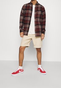 Chemise en flanelle à carreaux rouges et noirs, t-shirt blanc, shorts beiges, baskets Nike rouges sur des chaussettes blanches, se tenant devant un fond uni.