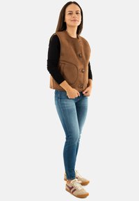 Gilet marrone senza maniche con scollo rotondo, chiusura a bottoni frontale e tasche laterali; abbinato a jeans blu e sneakers beige con accenti bordeaux.
