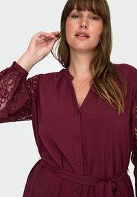 Burgundské šaty s zapínáním na knoflíky, výstřihem do V a dlouhými krajkovými rukávy. Materiál vypadá měkký s texturovaným povrchem. Nastavitelný detail zavazování v pase.