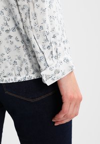 Blouse blanche à motif floral dans des tons bleus, avec des manches longues et des poignets boutonnés, associée à un jean en denim foncé.